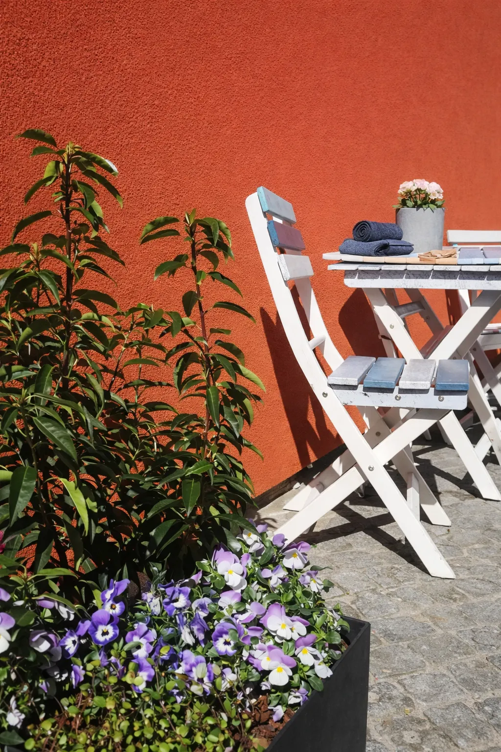 Detailaufnahme des Außenbereichs – weiße Klappstühle, Stiefmütterchen und Grünpflanzen vor orangefarbener Hauswand